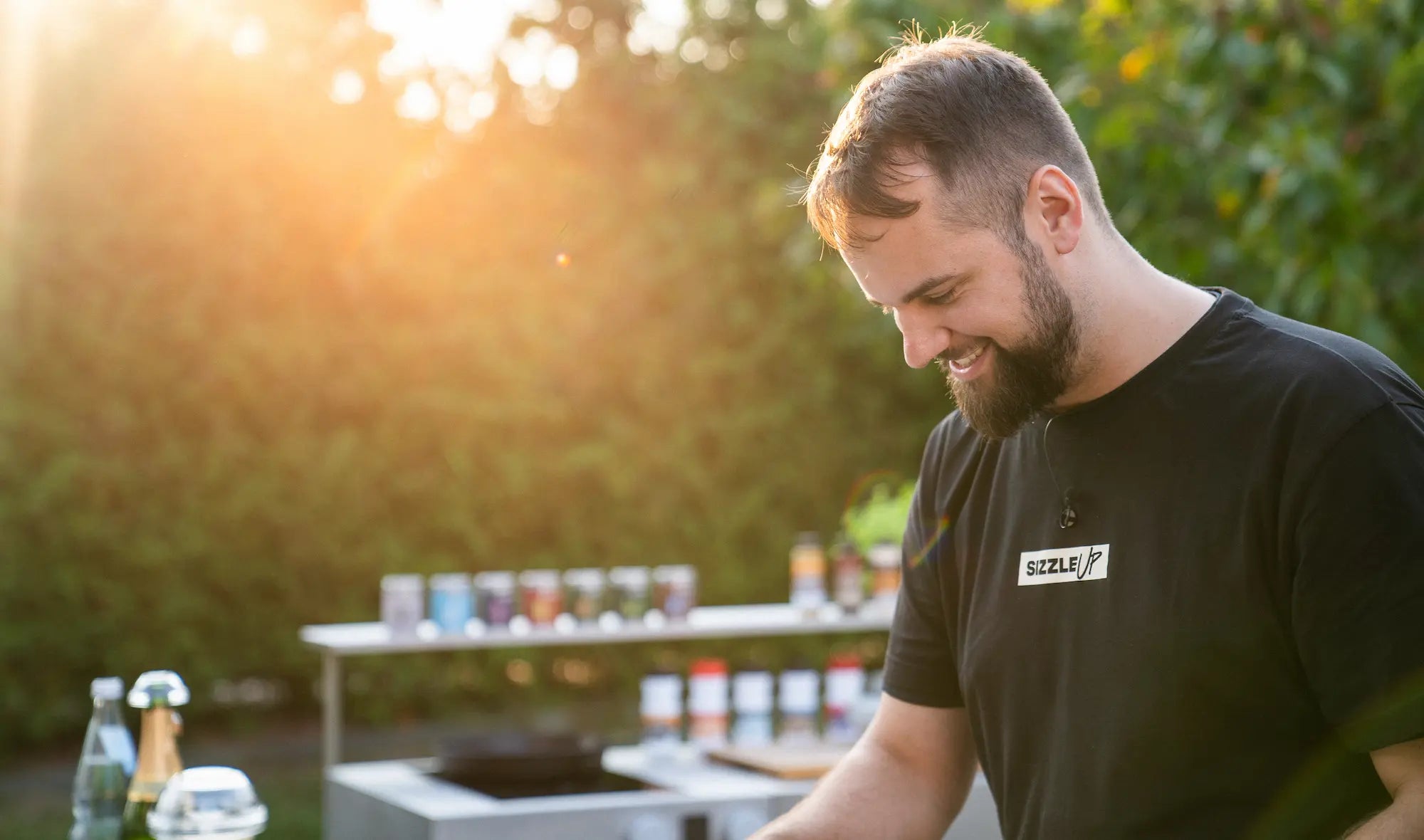 Ein lachender Mann mit Bart arbeitet im Freien an einem Grill, Sonnenuntergang im Hintergrund.