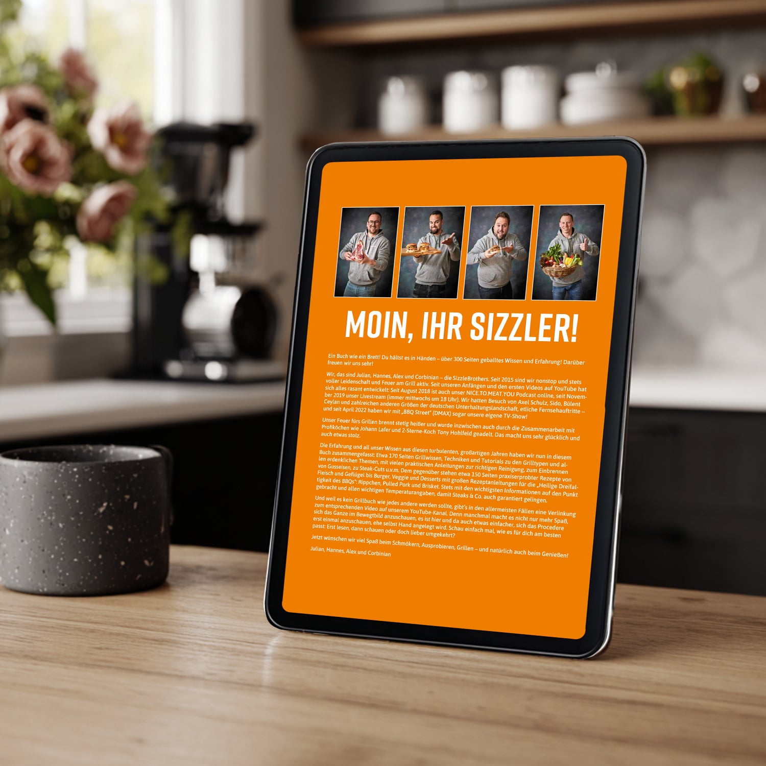 Tablet mit Begrüßungstext 'Moin, Ihr Sizzler!' auf orangefarbenem Hintergrund in einer Küche.