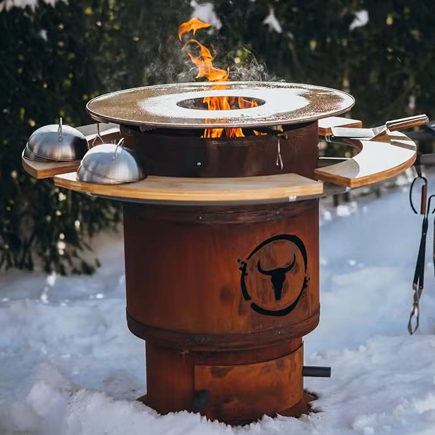 Rostiger Grill mit brennender Flamme im Schnee, umgeben von Bäumen.