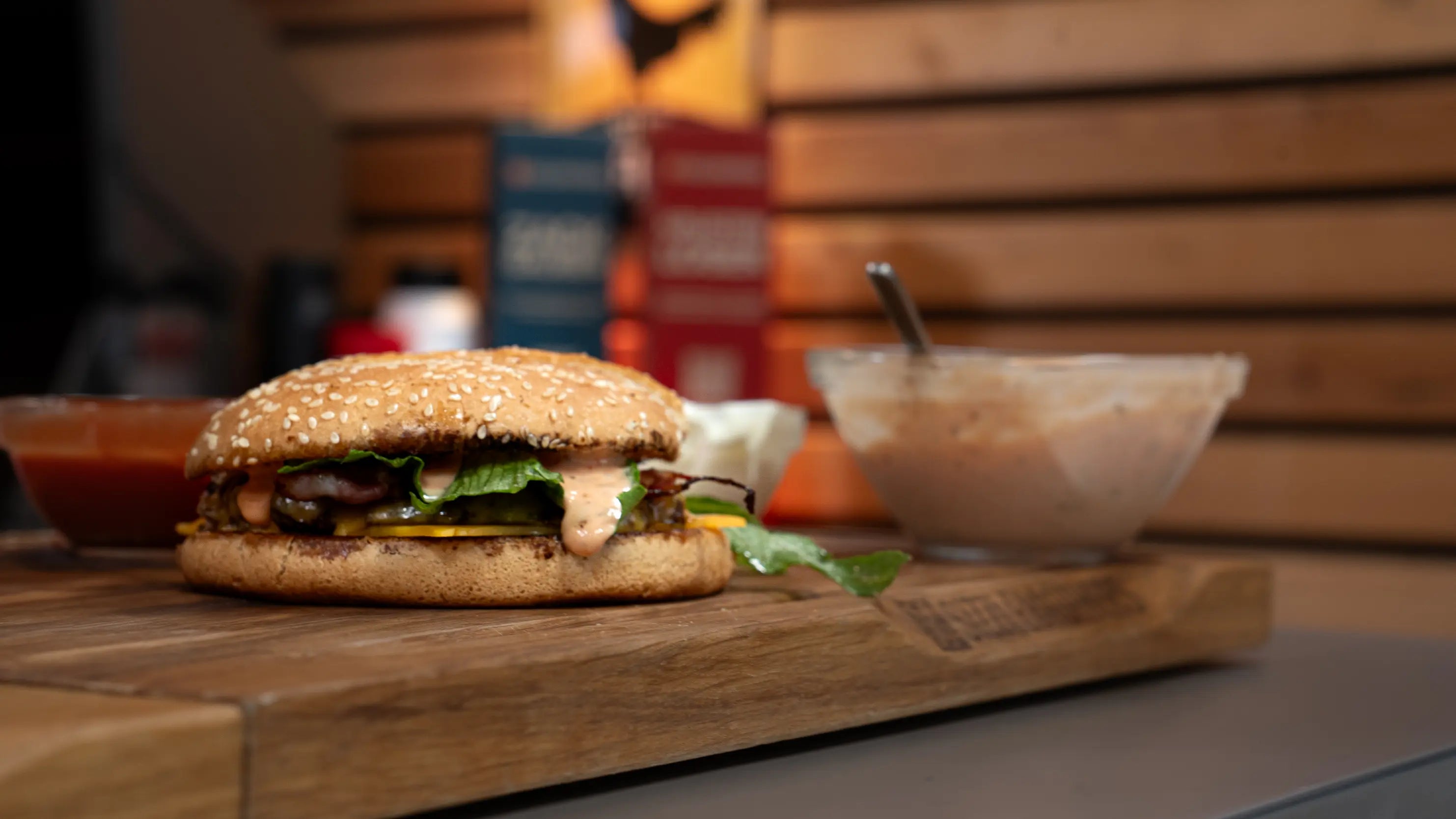 Burger mit Sesambrötchen, daneben eine Schüssel mit rosa Sauce auf Holzbrett.