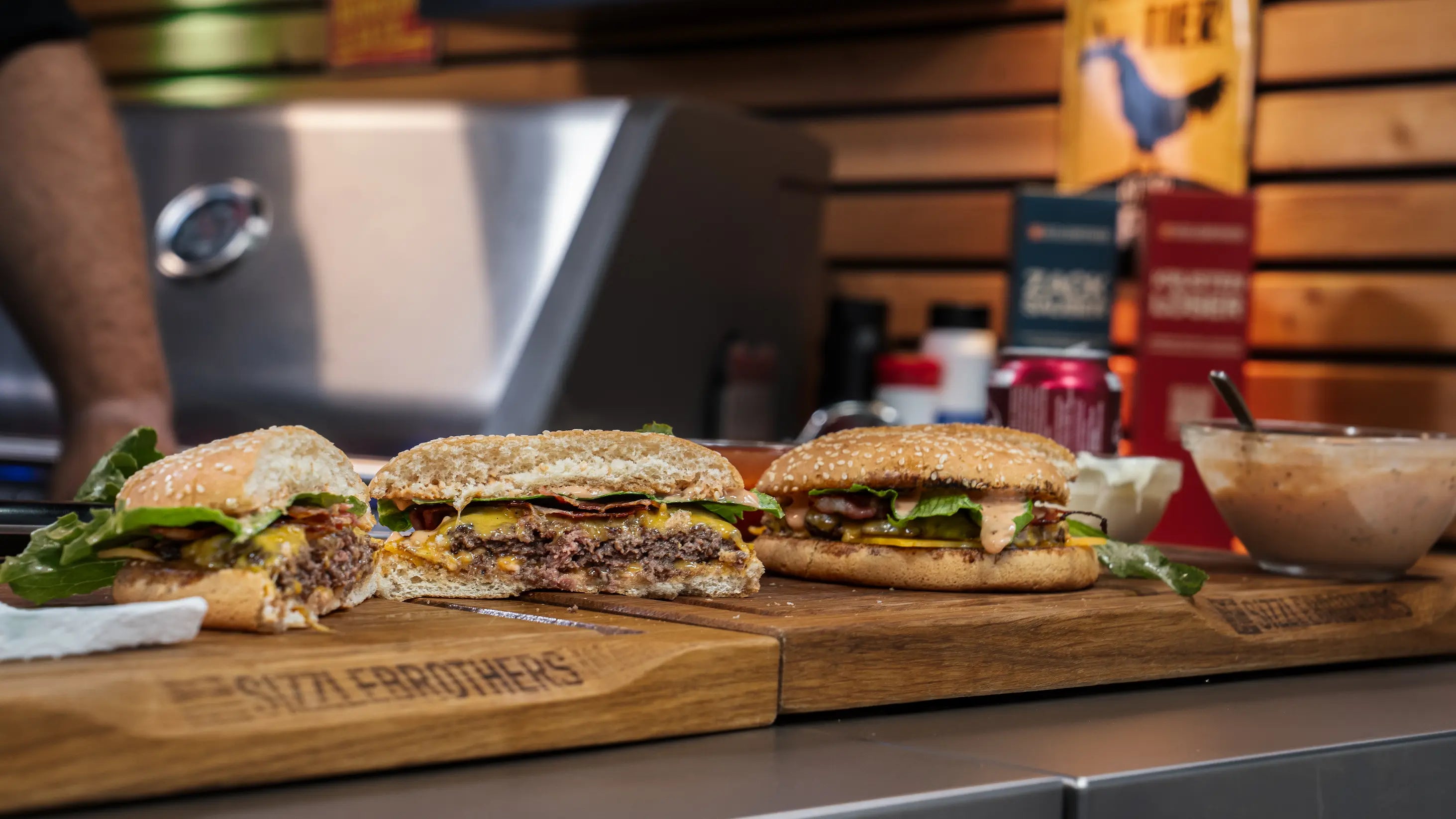 Zwei Burger mit Salat und Käse auf Schneidebrettern, dahinter Saucen.