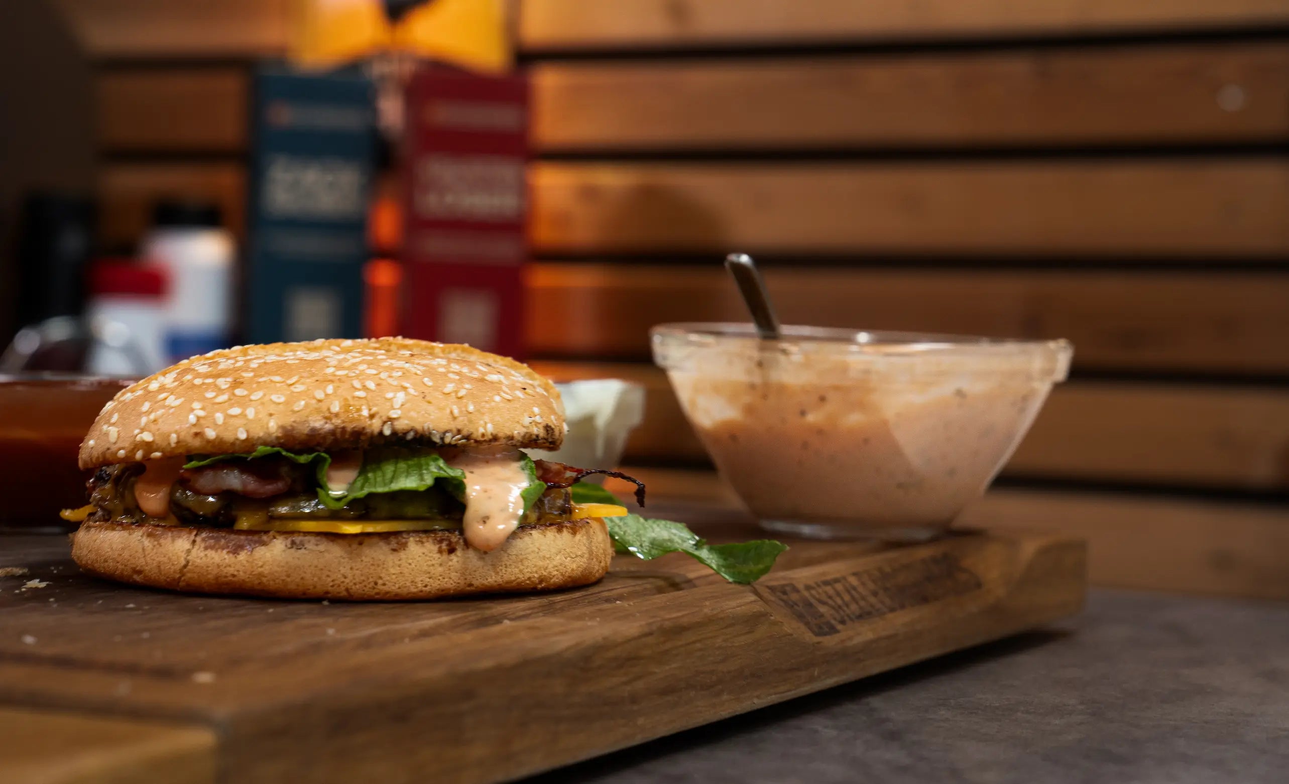 Burger mit Sesambrötchen und Salat auf einem Holzbrett neben einer Schüssel Sauce.