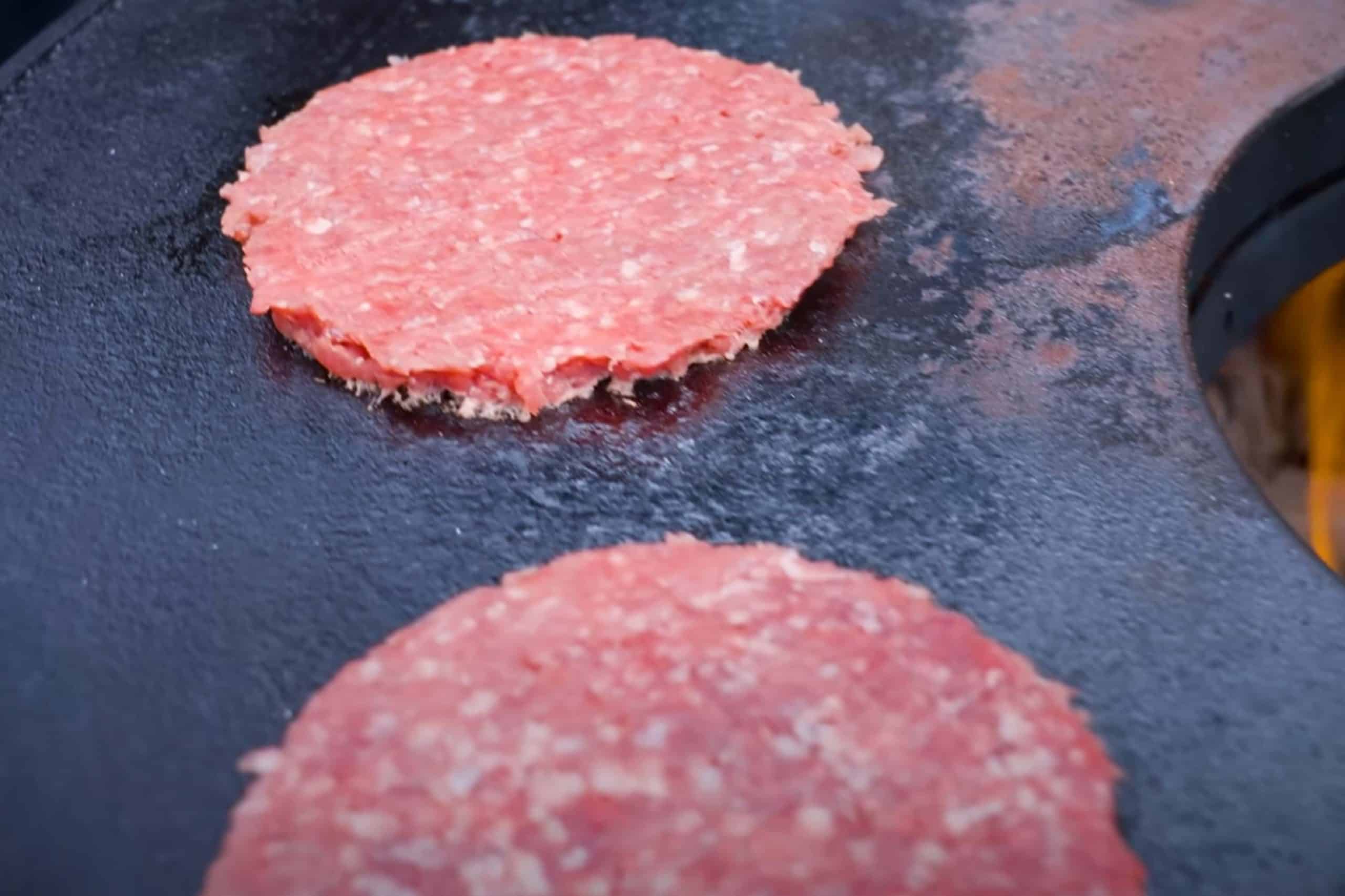 Zwei rohe Burger-Patties auf einer heißen, schwarzen Grillplatte.