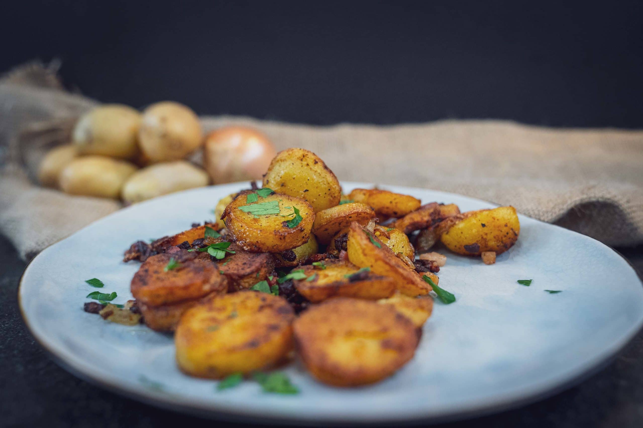 Gebratene Kartoffelscheiben mit Petersilie auf einem Teller vor einem Sack Kartoffeln.