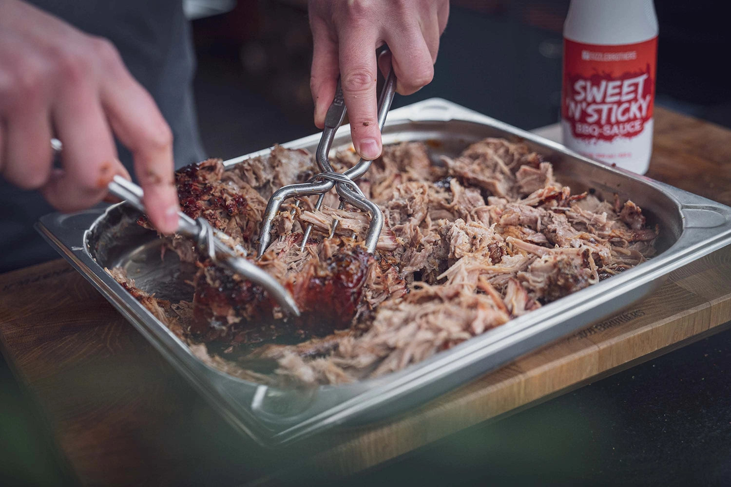 Person zerkleinert Braten in Metallschale, daneben BBQ-Sauce.