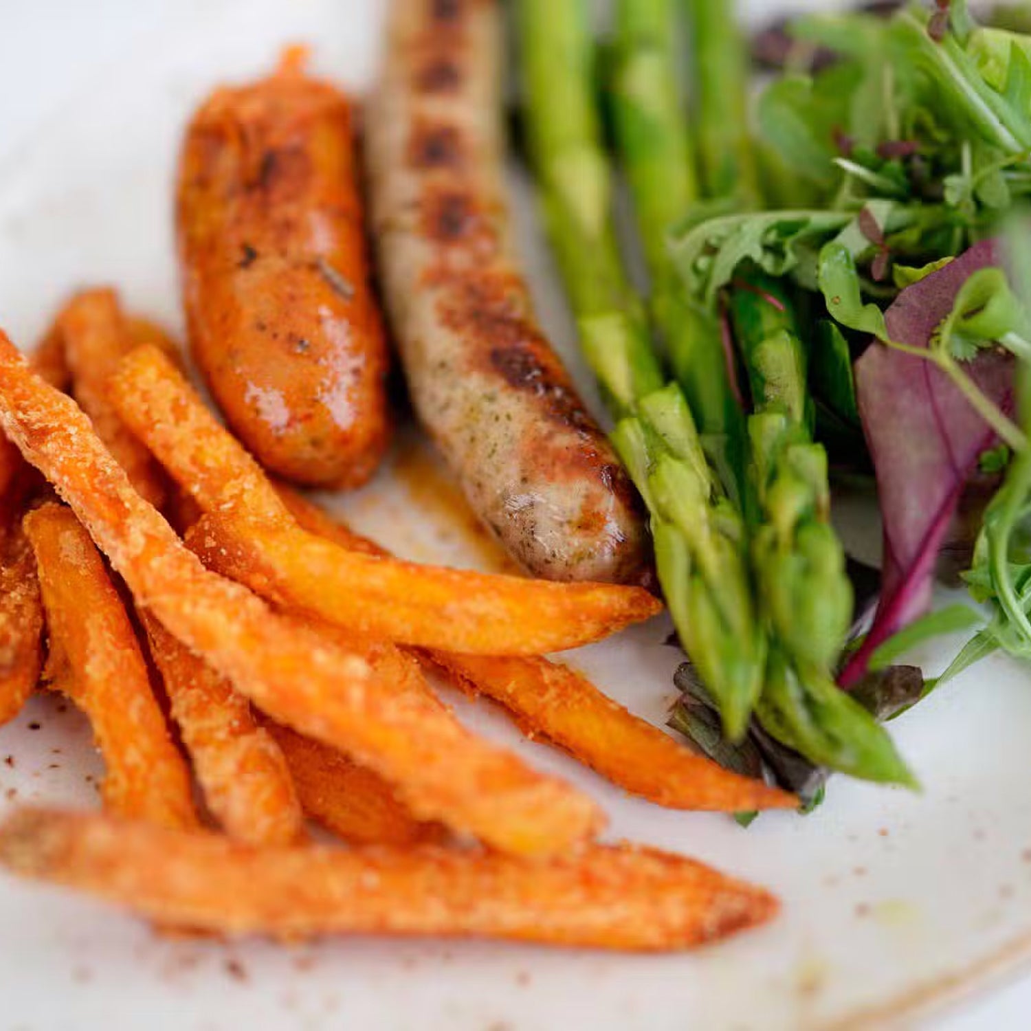 Teller mit Süßkartoffelpommes, Wurst, Spargel und Salat.
