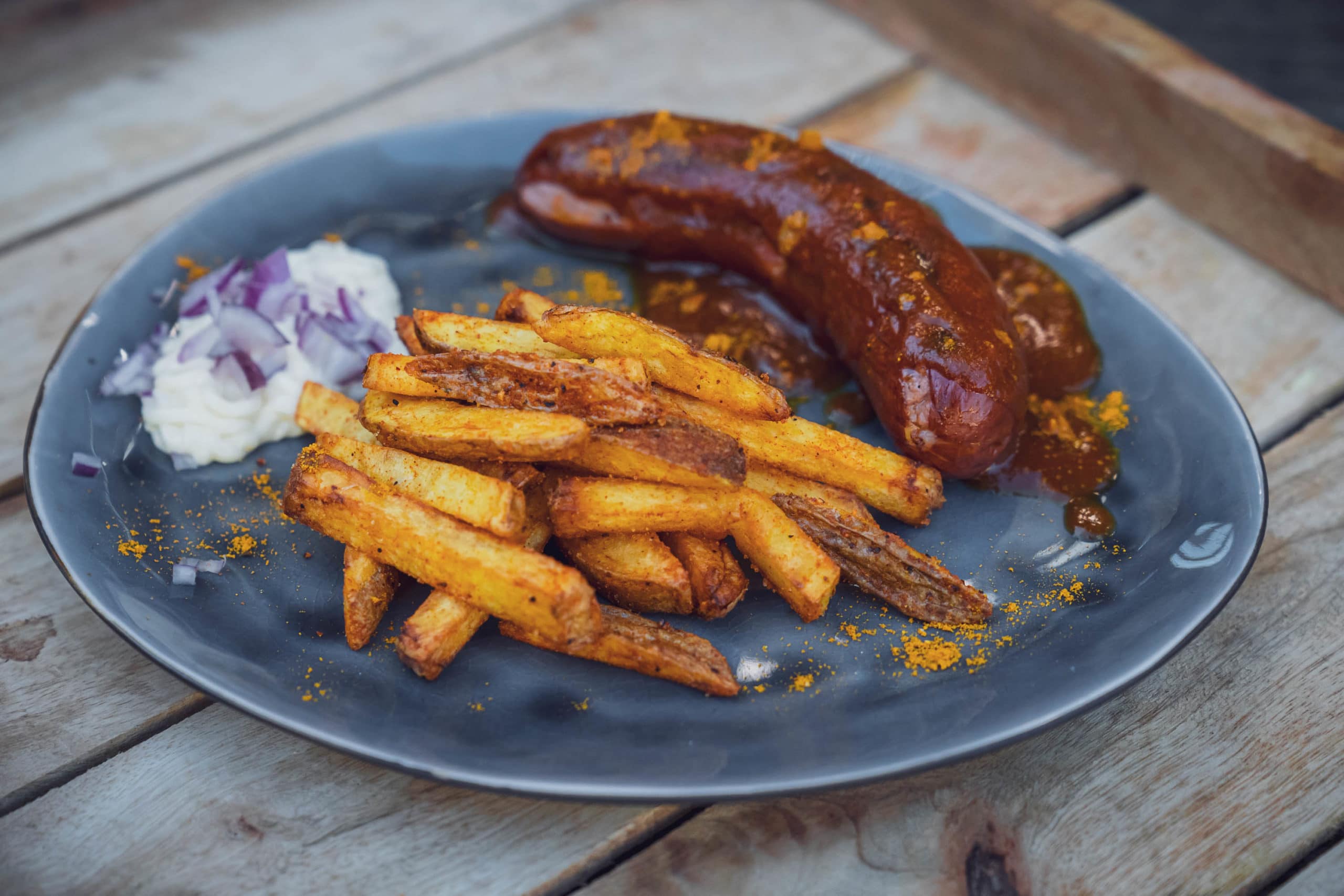 Currywurst mit Sauce, Pommes und Zwiebeln auf einem grauen Teller serviert.