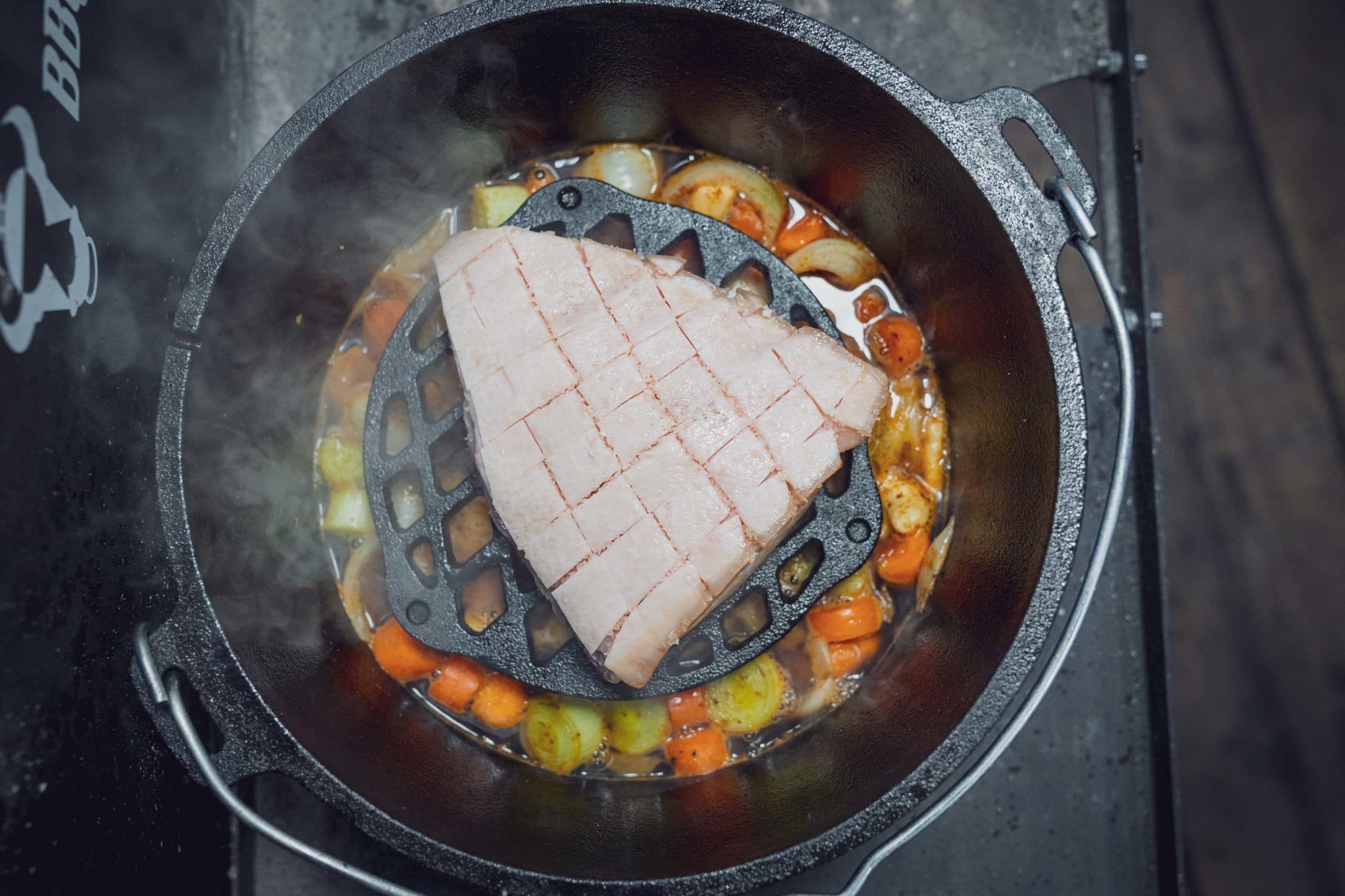 Ein Stück Fleisch auf einem Gitter, umgeben von Karotten und Kartoffeln im Topf.