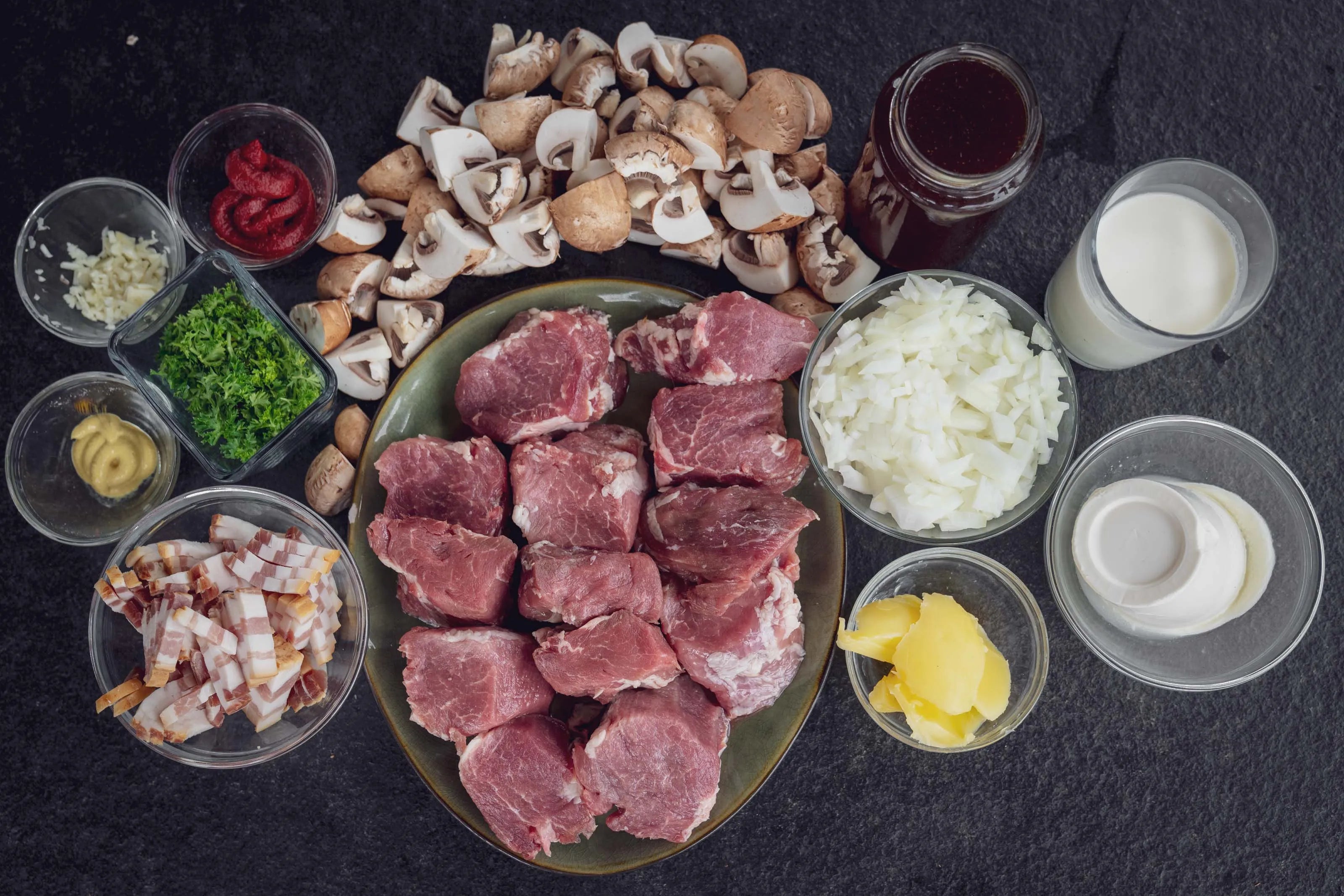 Teller mit rohem Fleisch und Schalen mit Zutaten auf schwarzem Hintergrund.