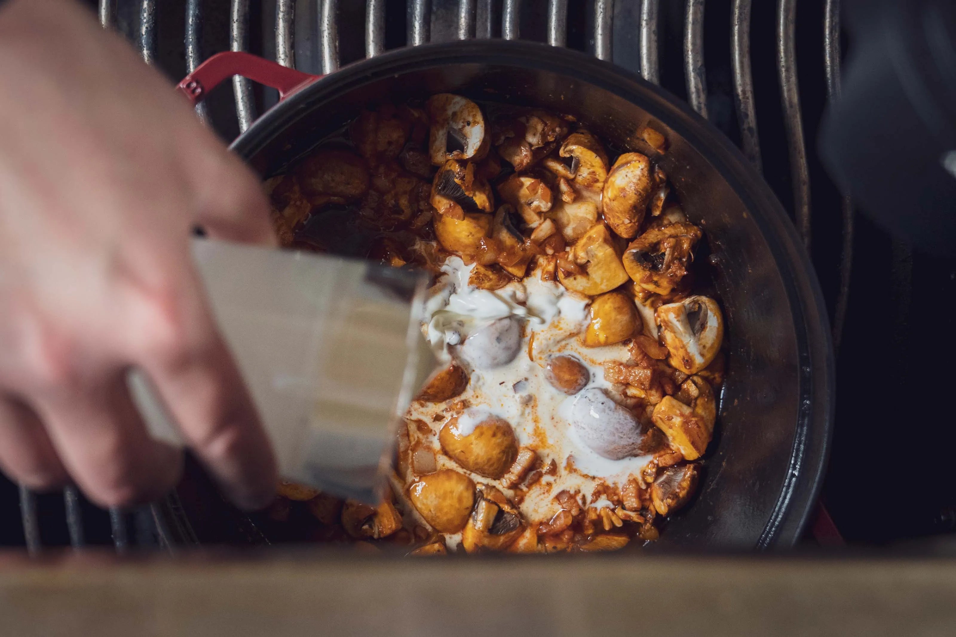 Hand gießt Sahne in einen Topf mit kochenden Pilzen.
