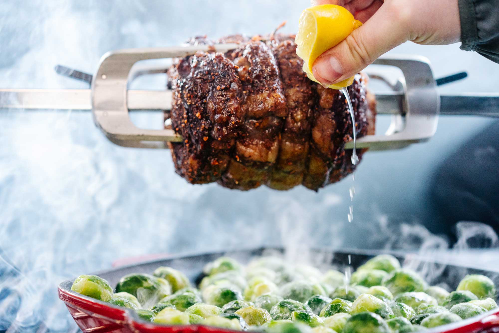 Zu sehen ist der Rib Eye Braten an einem Drehspieß und eine Zitrone, die ausgepresst wird. Der Saft tropft in die Pfanne mit dem Rosenkohl