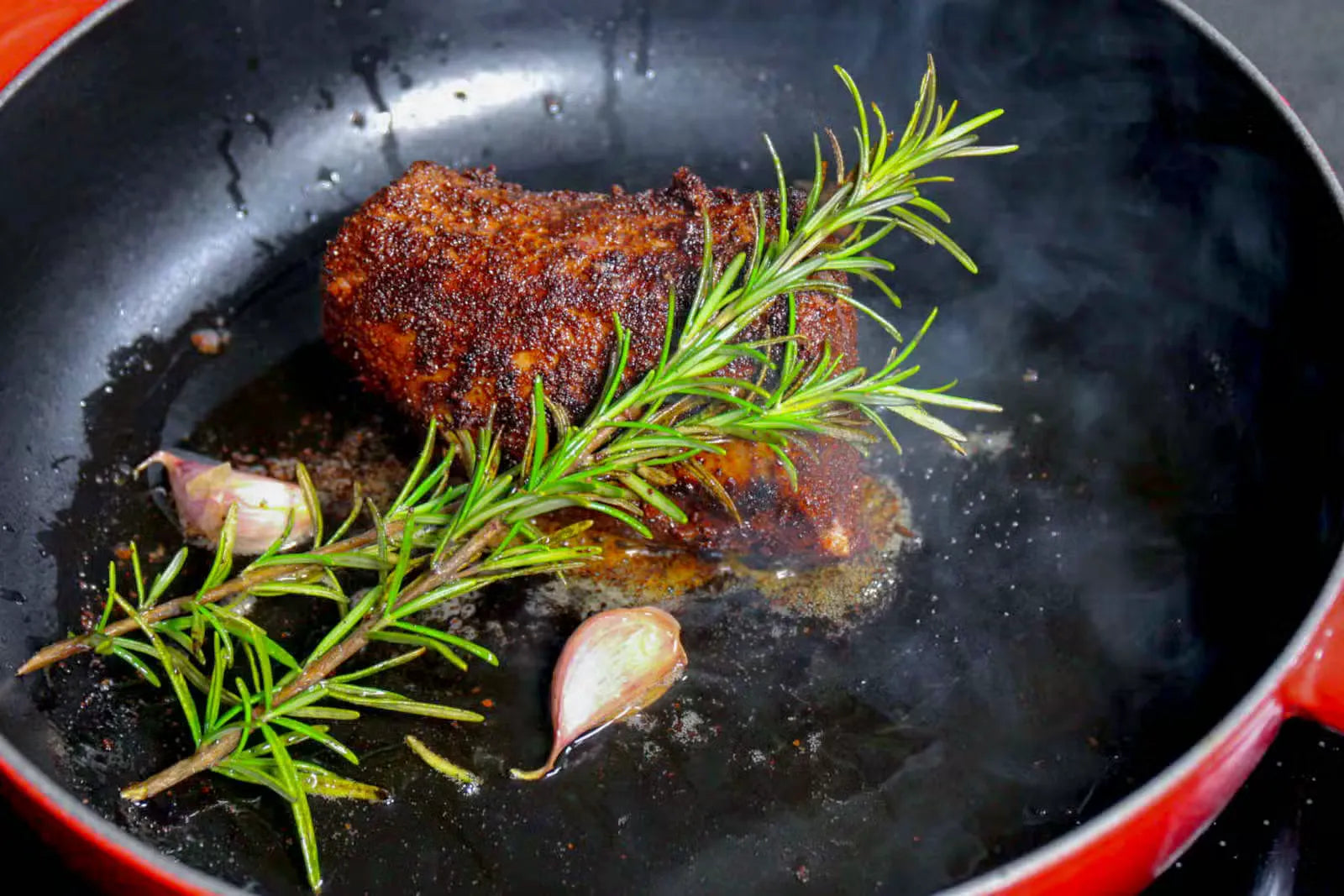 Ein Stück Fleisch mit Rosmarin und Knoblauch in einer schwarzen Pfanne.