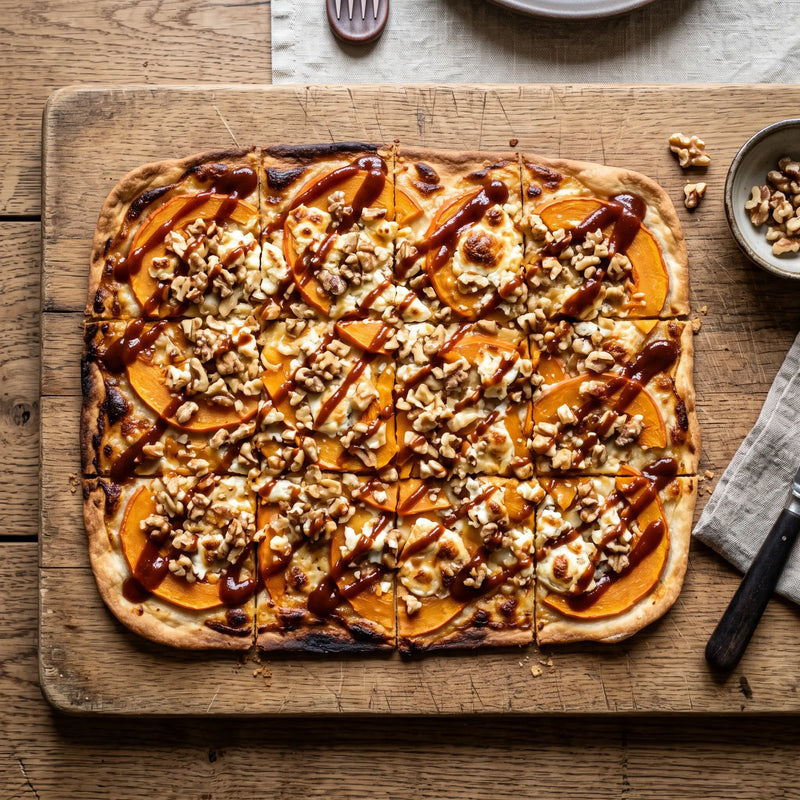 Rechteckiger Flammkuchen mit dünnem, kross gebackenem Teig, belegt mit dünnen Kürbisscheiben, Ziegenkäse, gehackten Walnüssen und brauner Sauce