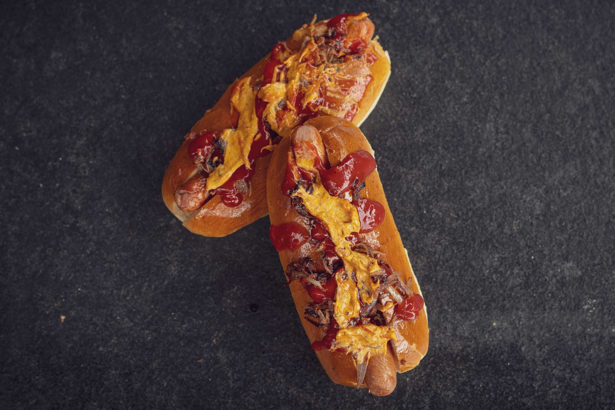 Zwei Hotdogs mit Ketchup und gelbem Käse auf dunklem Hintergrund.