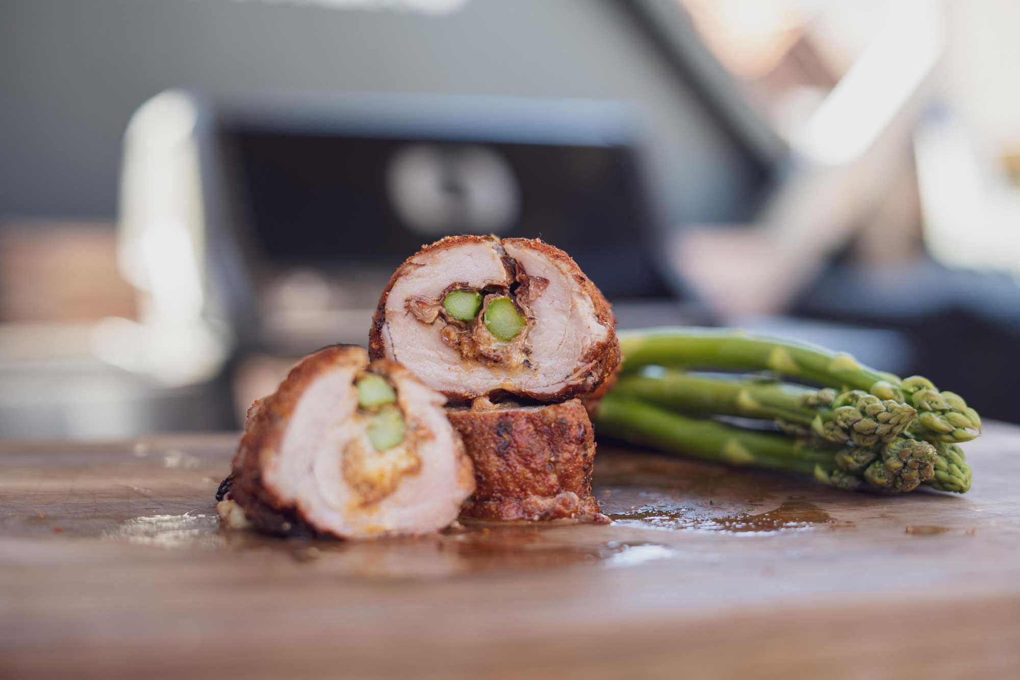 Gefüllte Hühnerbrust mit grünem Spargel auf Holzbrett, im Hintergrund Grill.