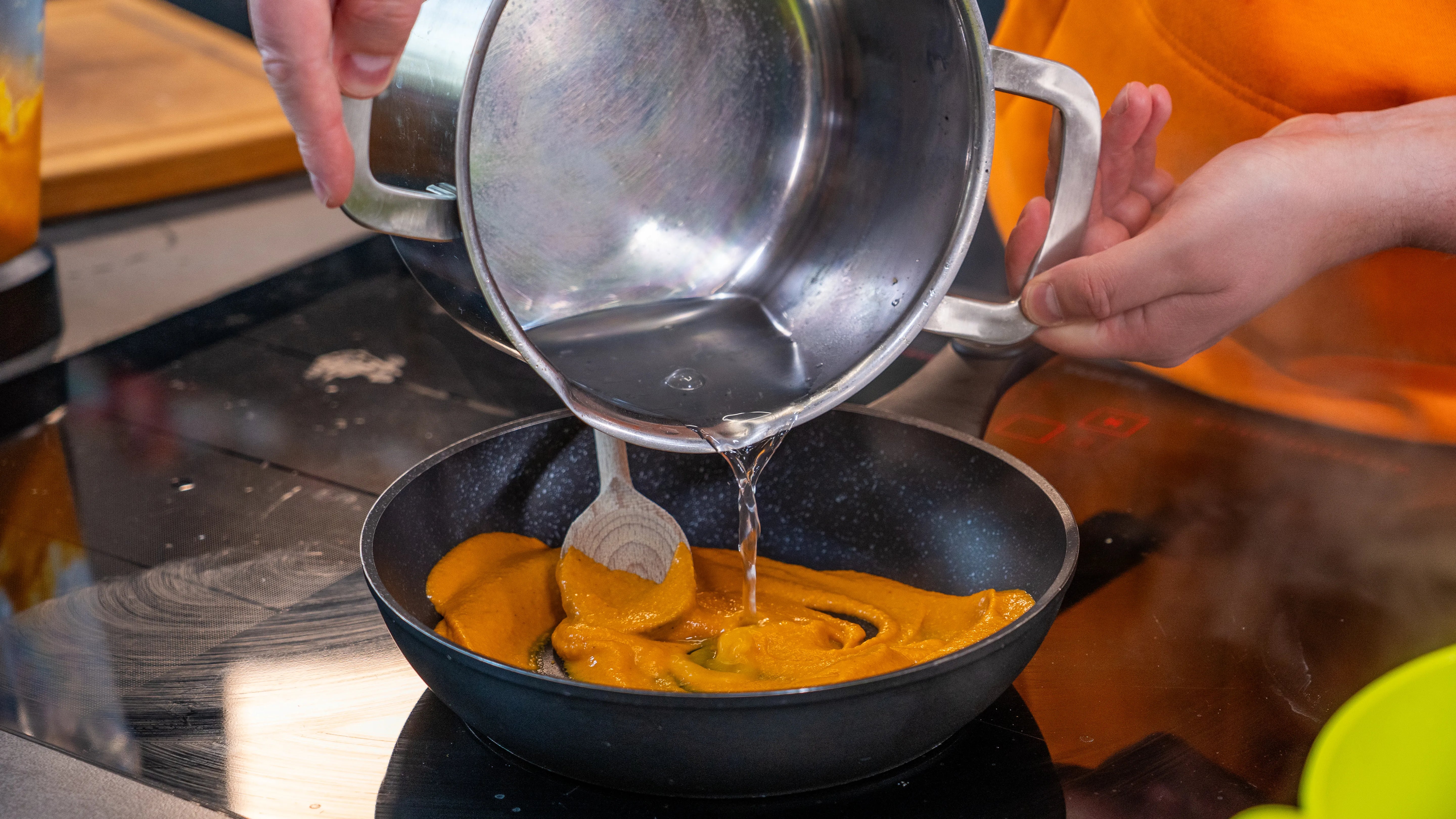 Hand gießt klare Flüssigkeit aus einem Topf in Pfanne mit orangener Soße.