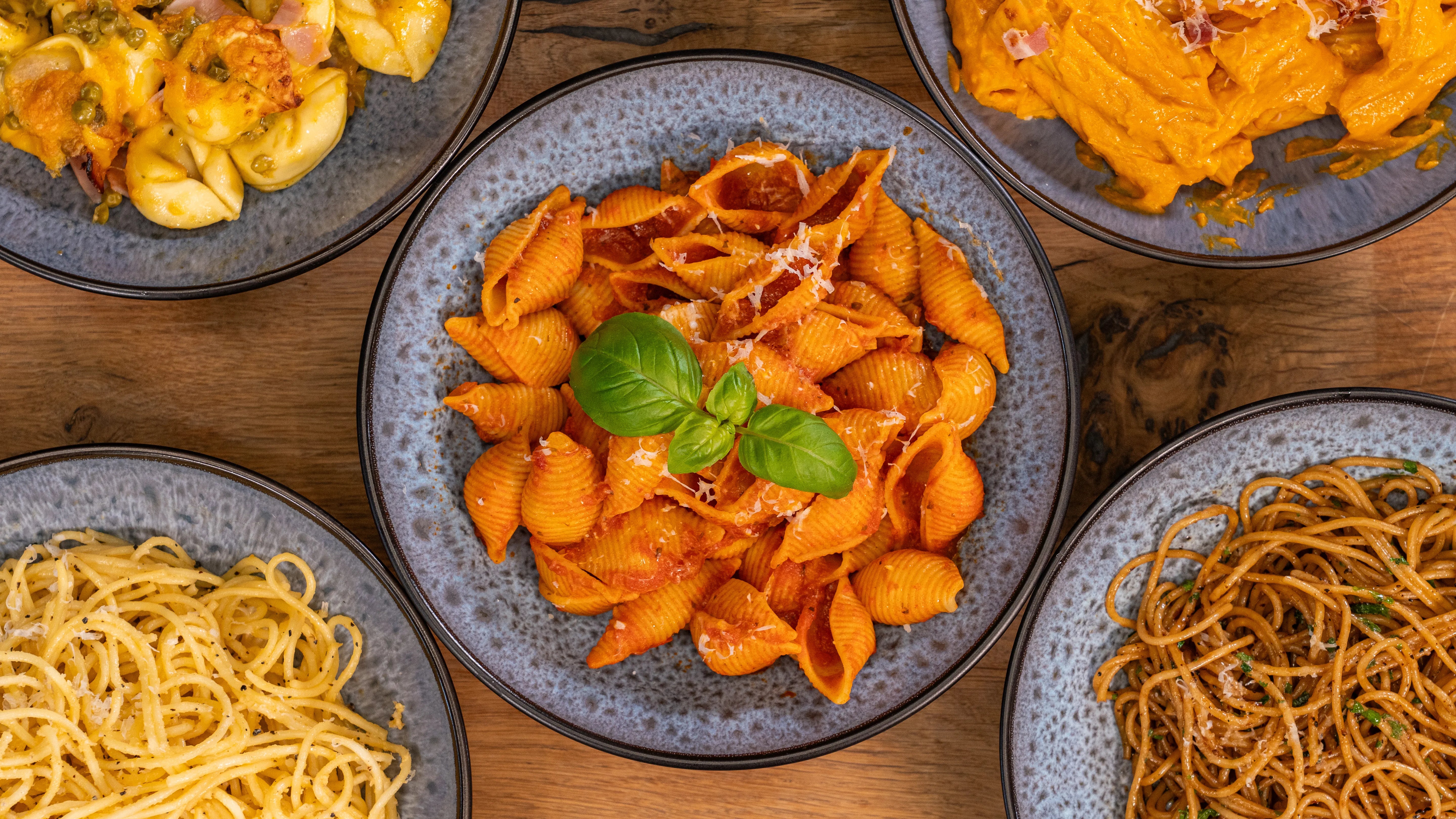 Fünf Pastagerichte in blauen Schüsseln auf Holzuntergrund drapiert.