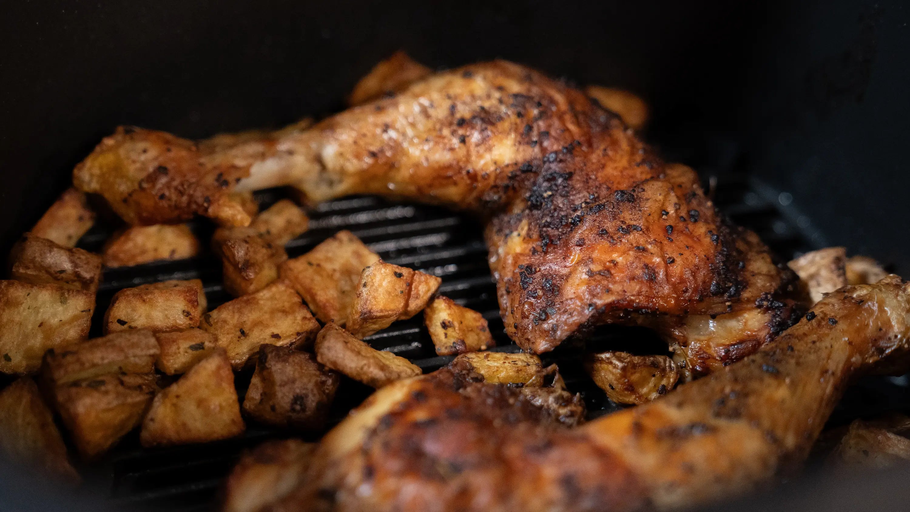 Knusprige Hähnchenkeulen und Kartoffelstücke in einem Airfryer.