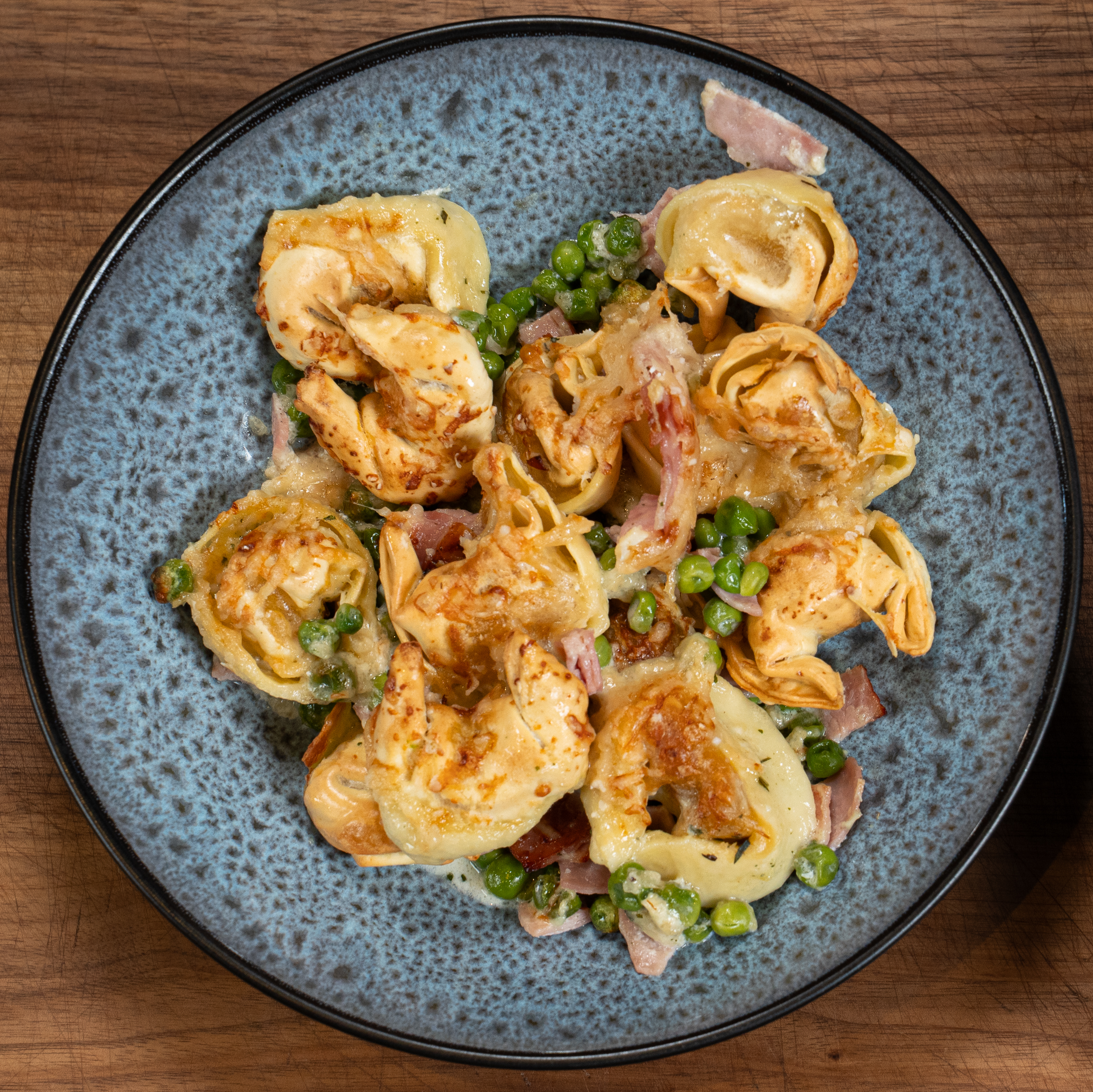 Tortellini mit Schinken und Erbsen auf einem blauen Teller.