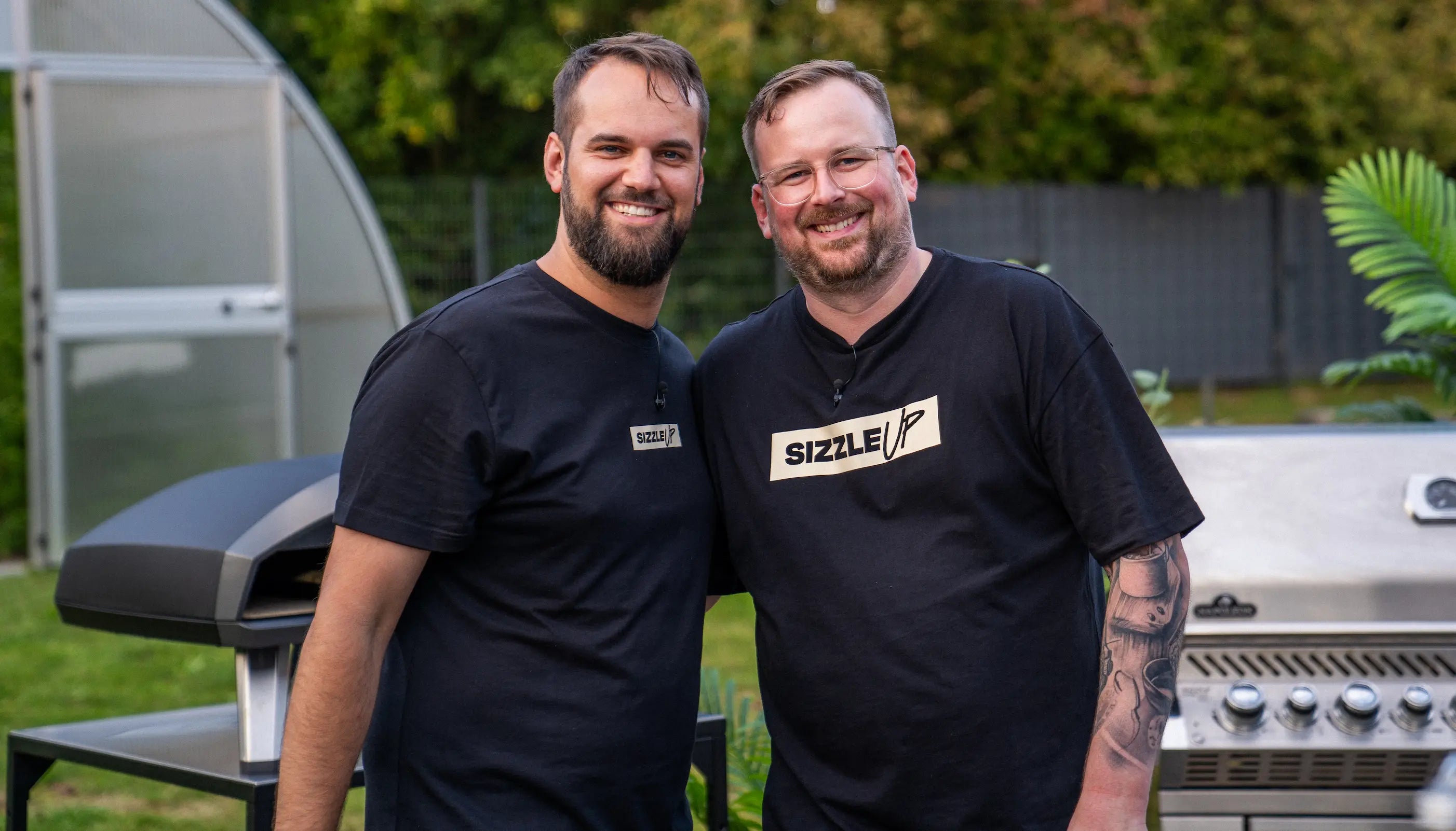 Zwei Männer in schwarzen T-Shirts stehen lächelnd vor einem Grill im Freien.
