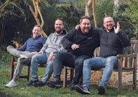 Vier lachende Männer sitzen auf Bänken im Garten.