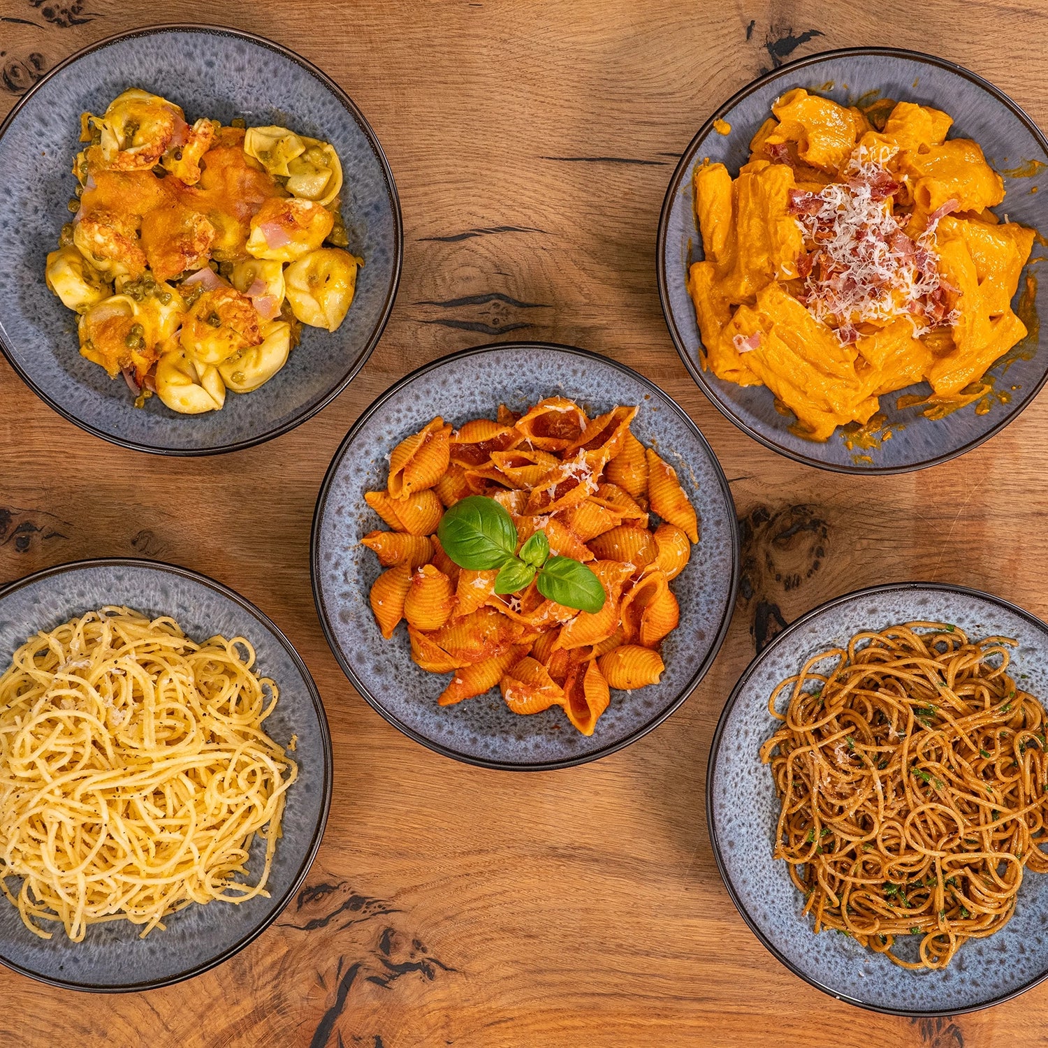 Fünf blaue Teller mit verschiedenen Pastagerichten auf einem Holztisch.
